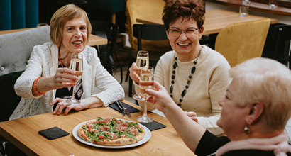 Three senior women toasting with glasses of sparkling drinks around a table with a pizza, celebrating community engagement and social connection at The Residence at Gramercy.